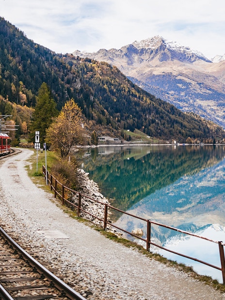 Red Bernina train traveling along a lake with mountain reflections near St. Moritz.