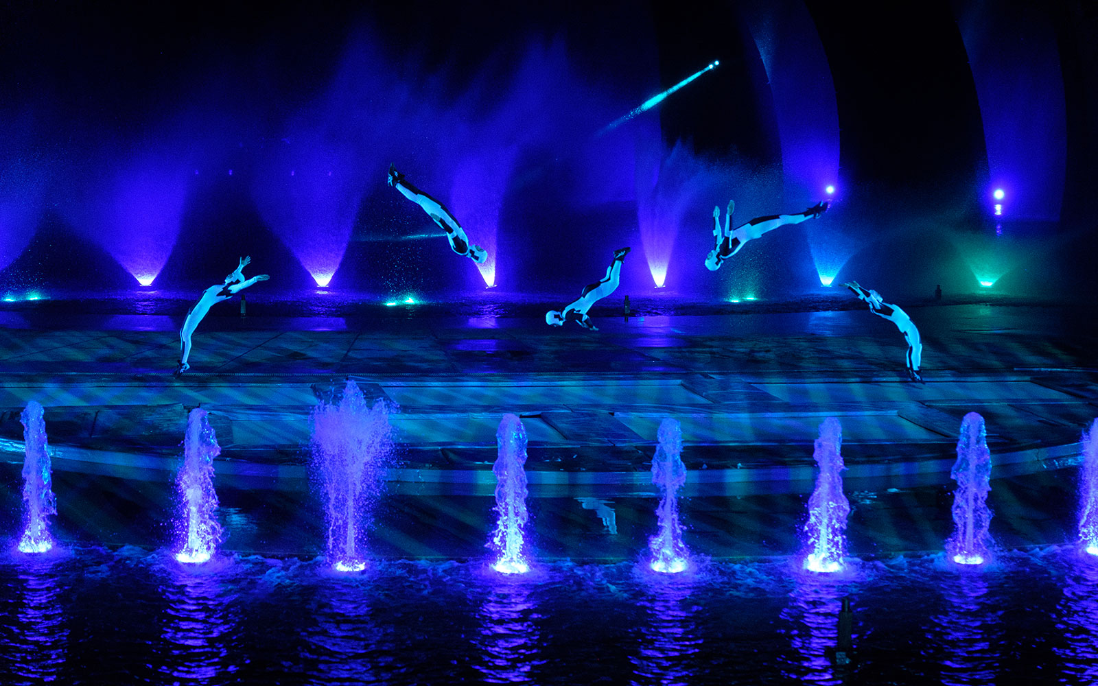 Acrobats performing mid-air flips over illuminated fountains at the Kiss of the Sea Show.
