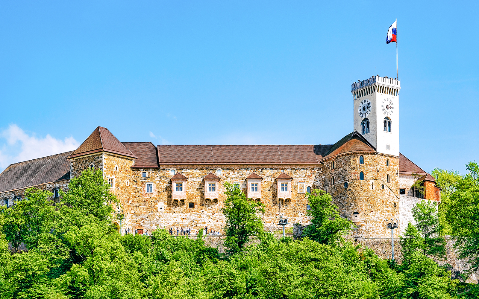 Ljubljana Castle with clock tower and Slovenian flag, surrounded by greenery.