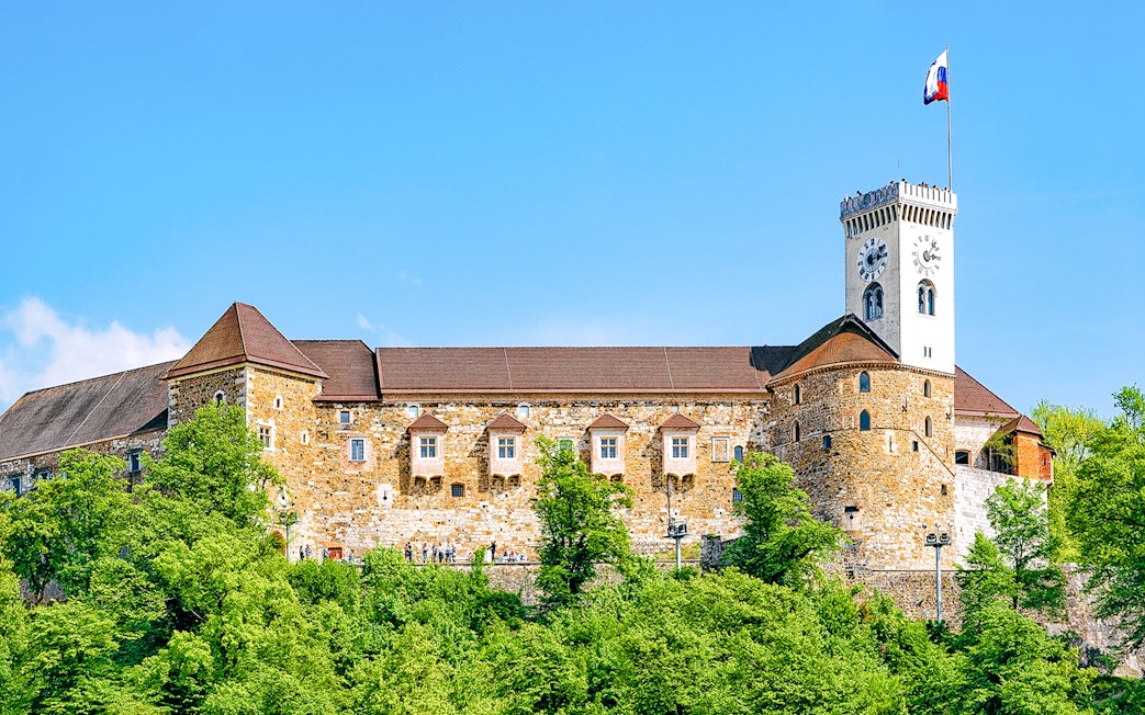 Ljubljana Castle with clock tower and Slovenian flag, surrounded by greenery.