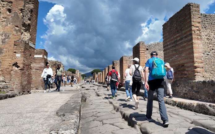 Tourists walking through ancient streets of Pompeii, Italy, with ruins and cloudy sky.