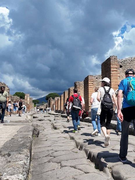Tourists walking through ancient streets of Pompeii, Italy, with ruins and cloudy sky.