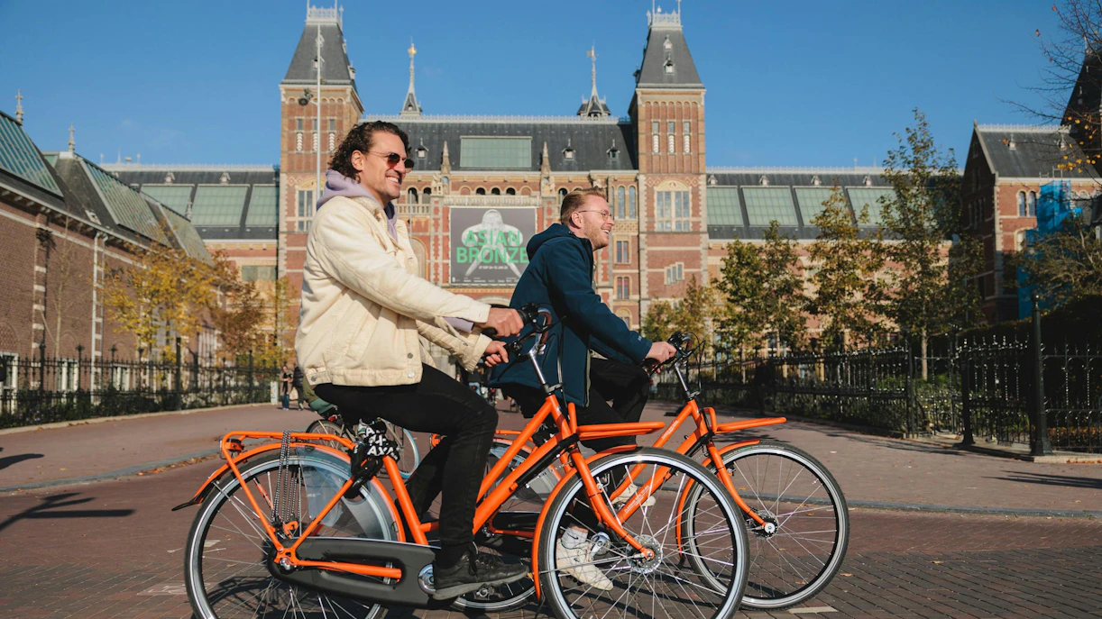 Cyclists on orange bikes in front of the Rijksmuseum during a city bike tour.