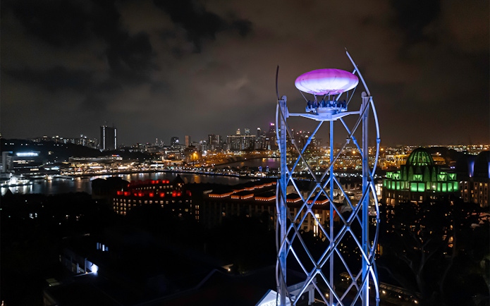 SkyHelix Sentosa ride illuminated at night with Singapore cityscape in the background.