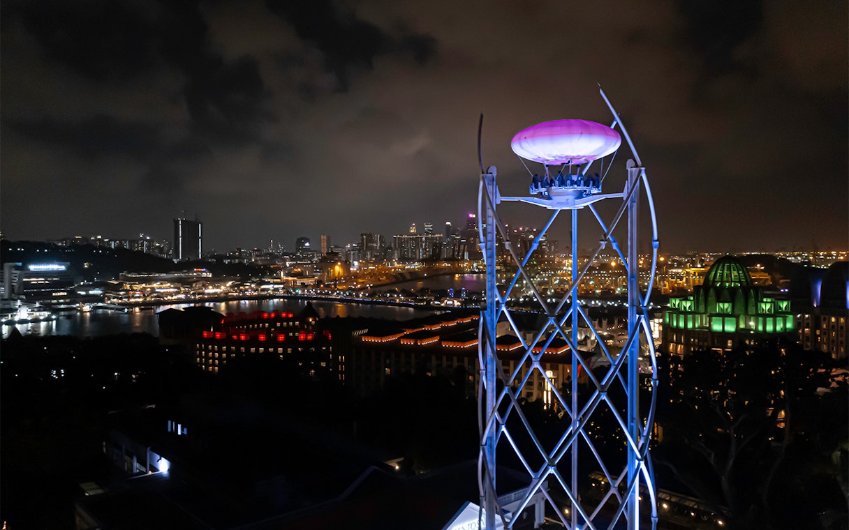 SkyHelix Sentosa ride illuminated at night with Singapore cityscape in the background.