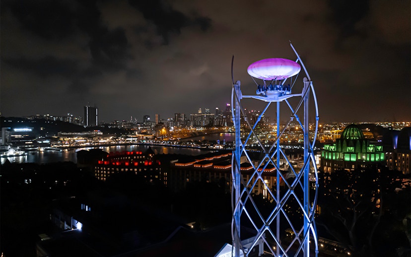 SkyHelix Sentosa ride illuminated at night with Singapore cityscape in the background.