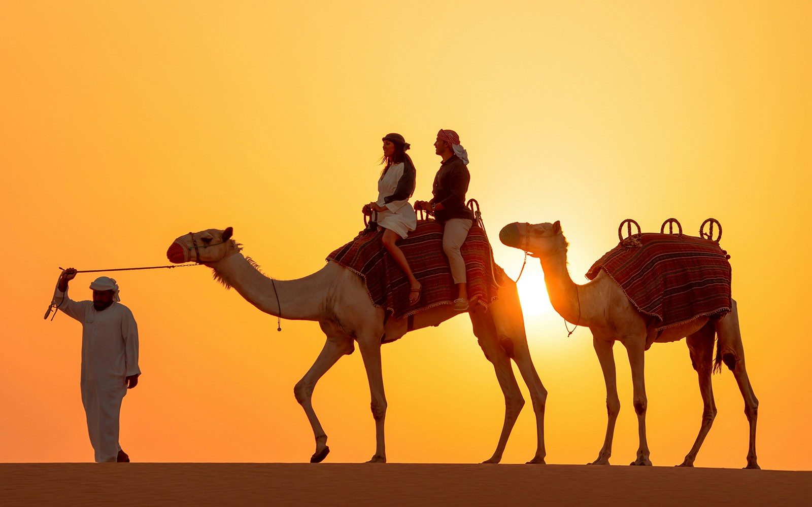 Tourists on camels led by a guide in the Dubai desert at sunset.