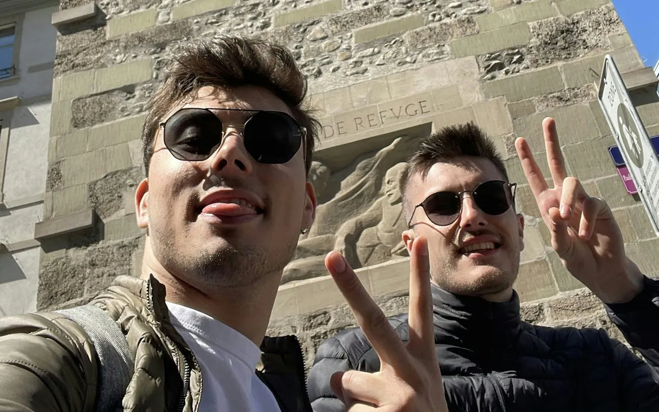Two people posing with peace signs in front of a historic stone relief in Geneva.