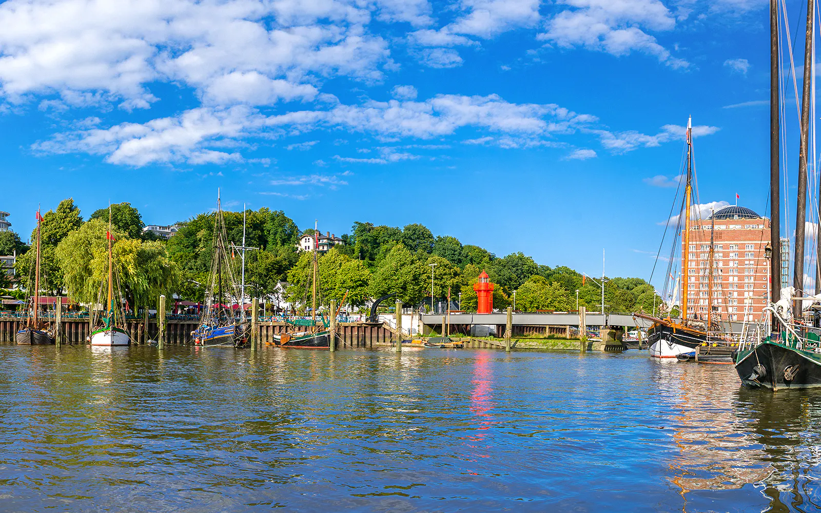 Boats docked along the Hamburg harbor with a red lighthouse and greenery in the background.