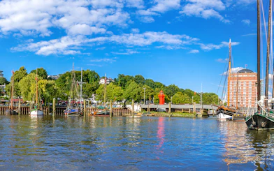 Boats docked along the Hamburg harbor with a red lighthouse and greenery in the background.
