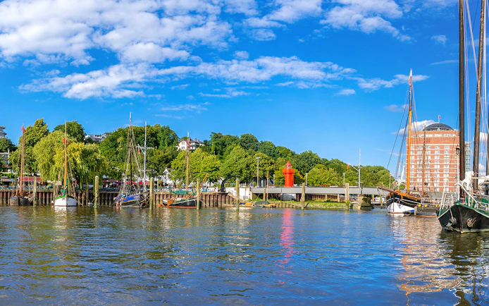 Boats docked along the Hamburg harbor with a red lighthouse and greenery in the background.