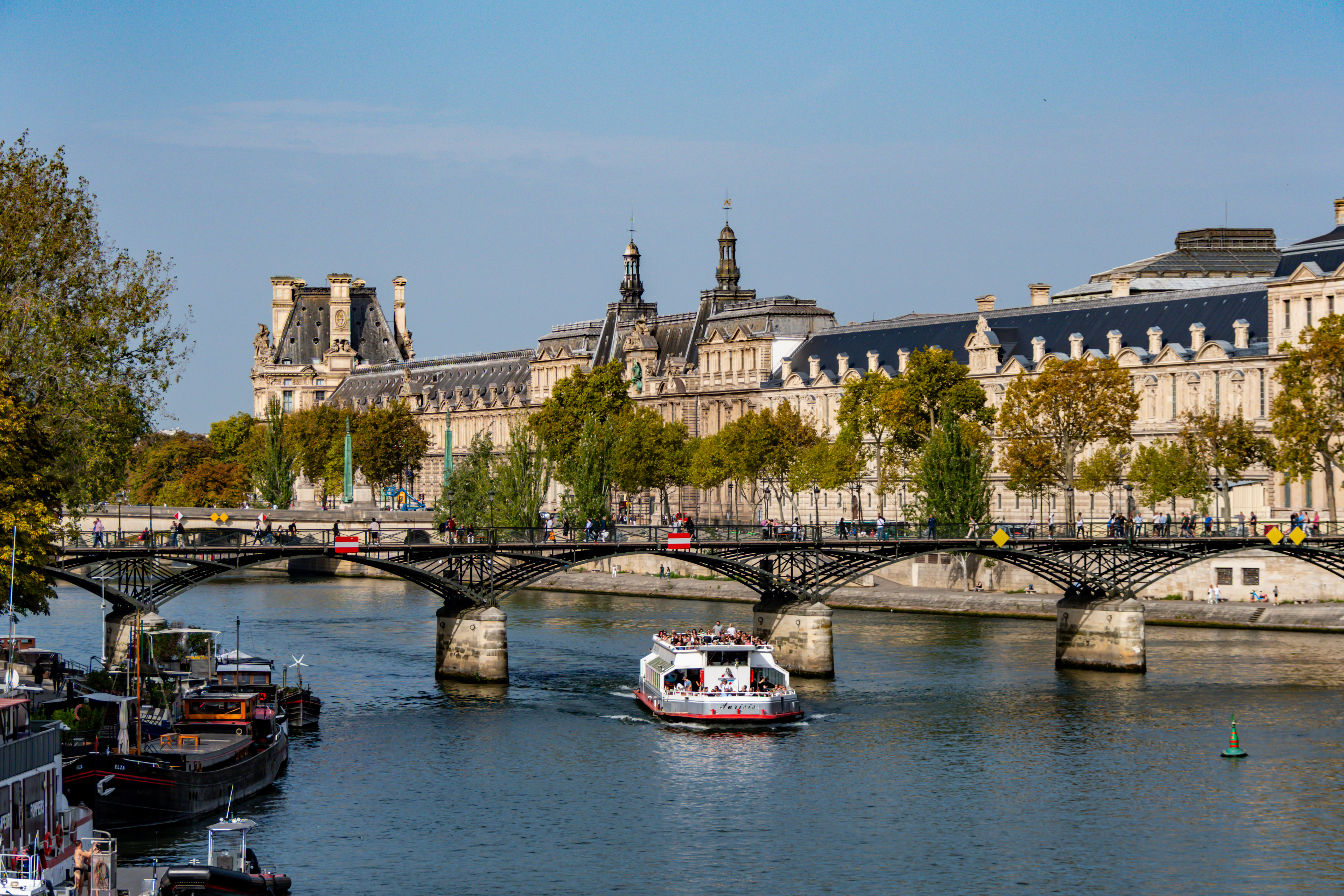 Seine River cruise boat passing under Pont des Arts with Louvre Museum in the background, Paris.