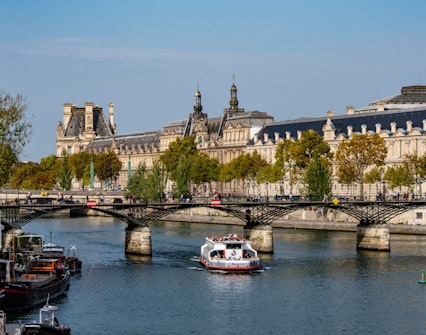 Seine River cruise boat passing under Pont des Arts with Louvre Museum in the background, Paris.