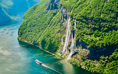 Seven Sisters waterfall cascading down cliffs in Geirangerfjord, Norway, with a boat in the fjord.