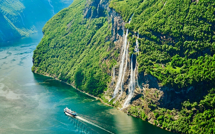 Seven Sisters waterfall cascading down cliffs in Geirangerfjord, Norway, with a boat in the fjord.