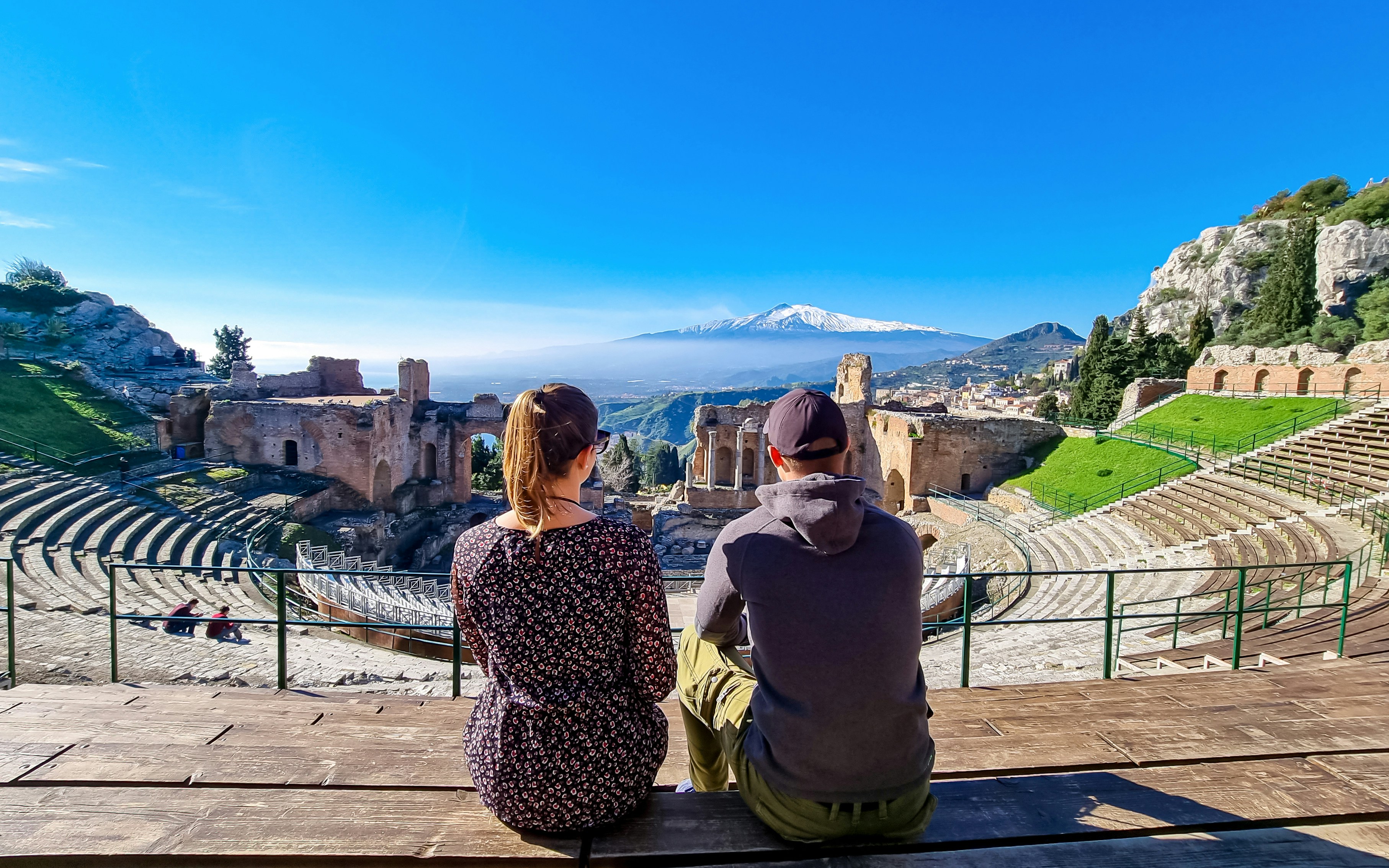 couple sitting with panoramic view on snow capped Mount Etna volcano on a sunny day seen from ancient Greek theater of Taormina, island Sicily, Italy