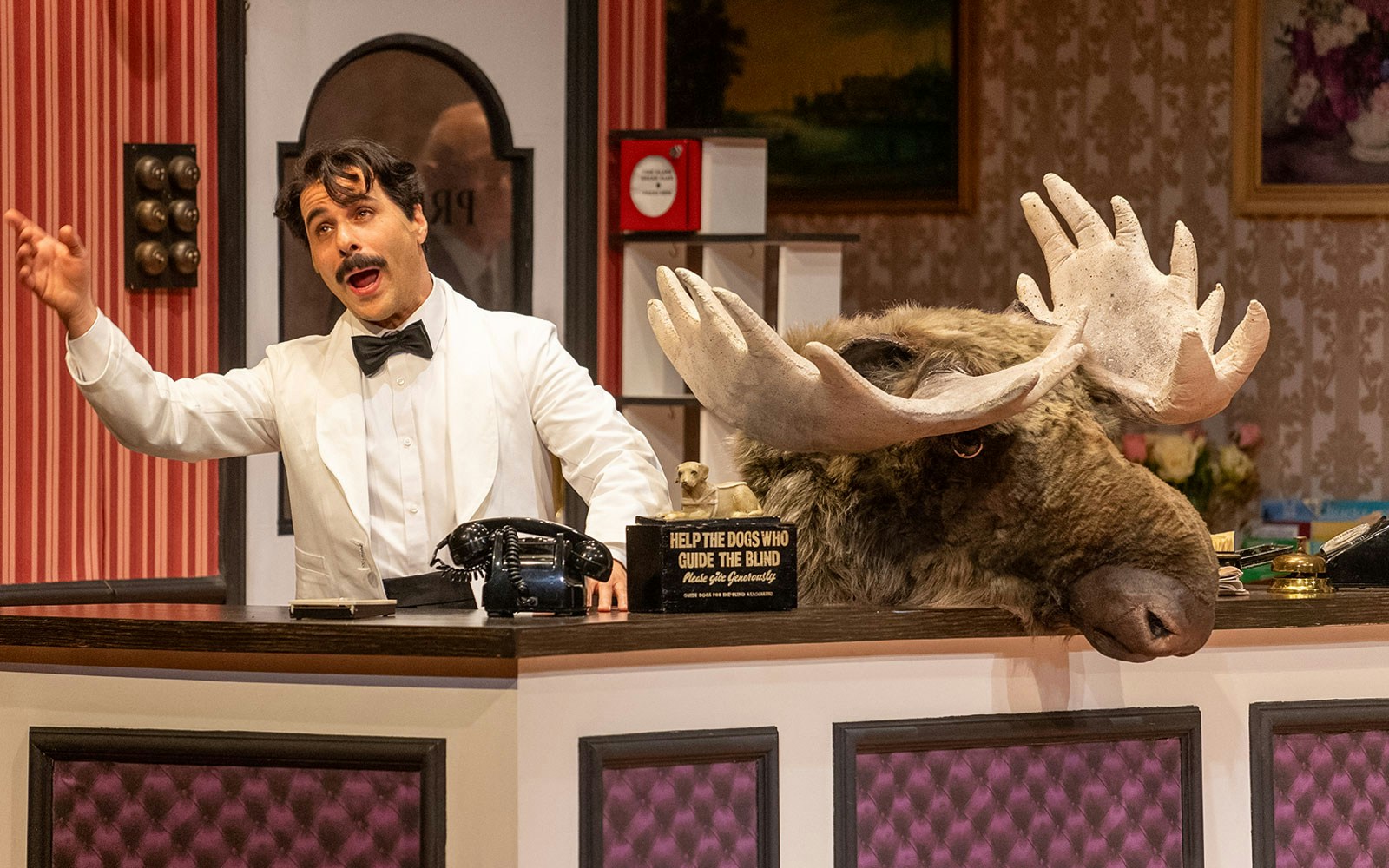 Hotel concierge in white tuxedo gestures beside a moose head at Fawlty Towers reception desk.