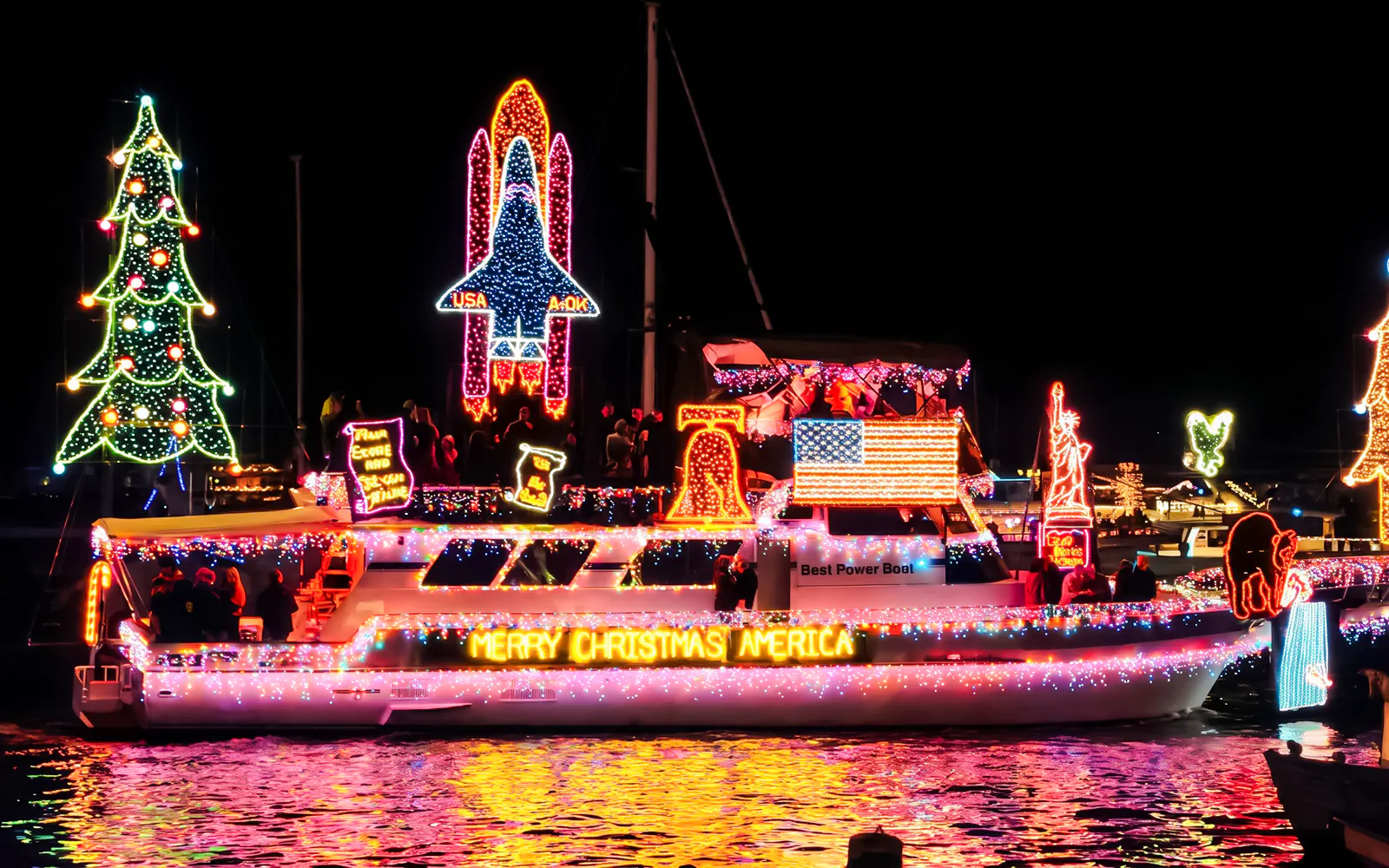 Christmas boat decorated with lights, featuring a tree and space shuttle, during holiday parade.