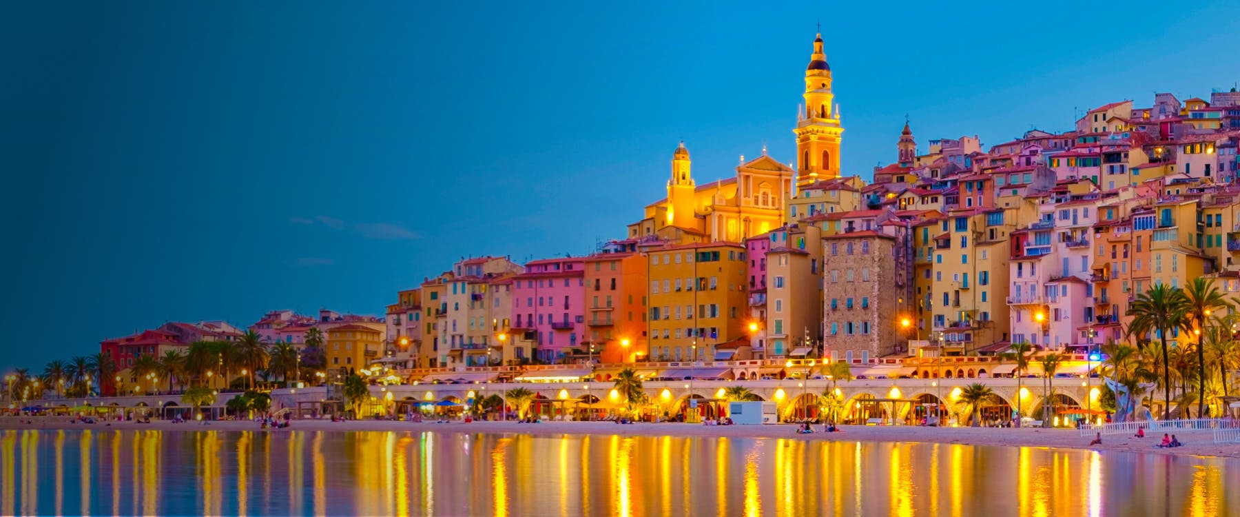 Colorful buildings and illuminated church in Menton, Provence, reflecting on the water at dusk.