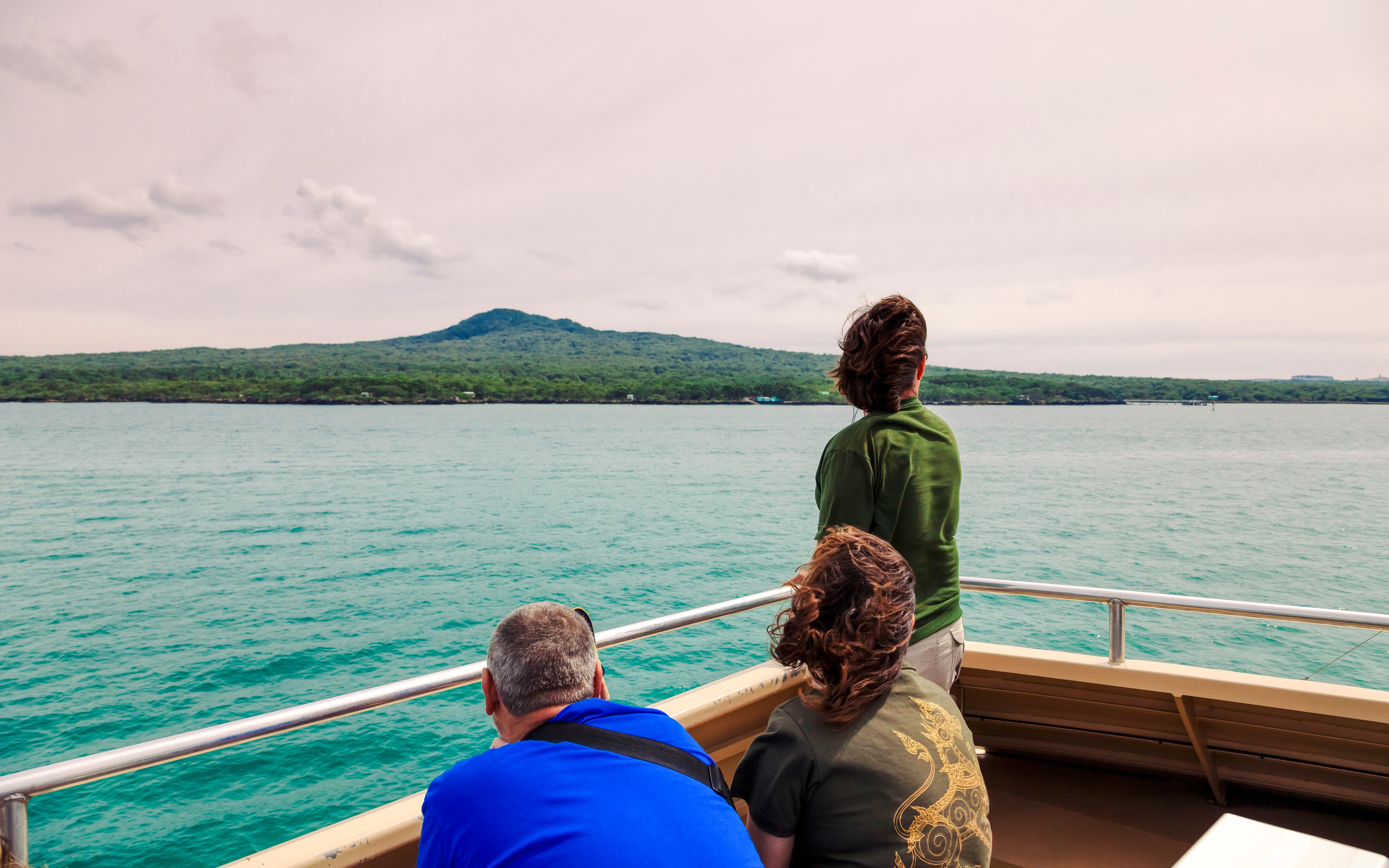 Tourists on a ferry approaching Rangitoto Island, Auckland, with lush greenery and volcanic landscape.