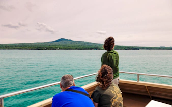 Tourists on a ferry approaching Rangitoto Island, Auckland, with lush greenery and volcanic landscape.