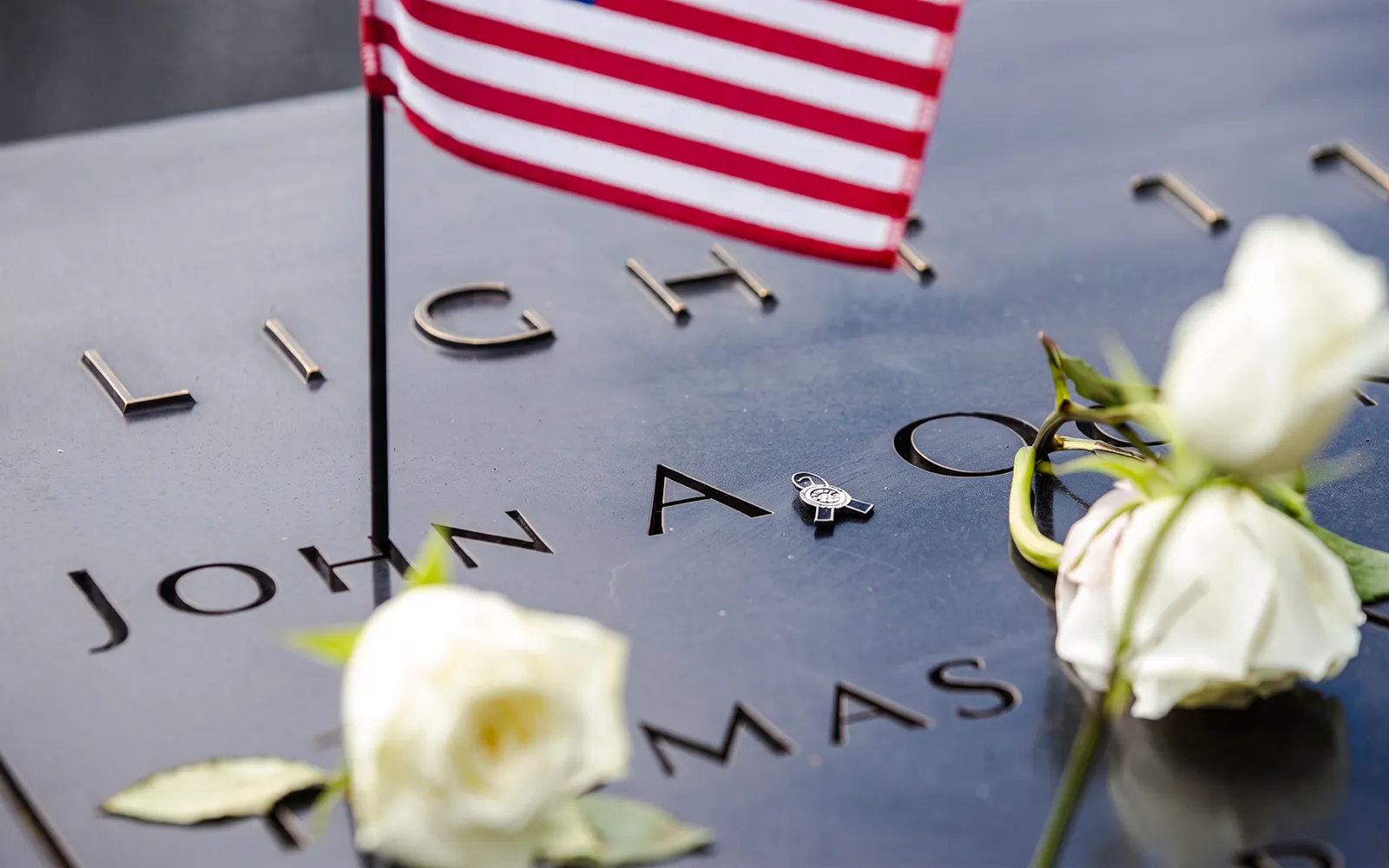 Names engraved on the 9/11 Memorial with white roses and a small American flag.