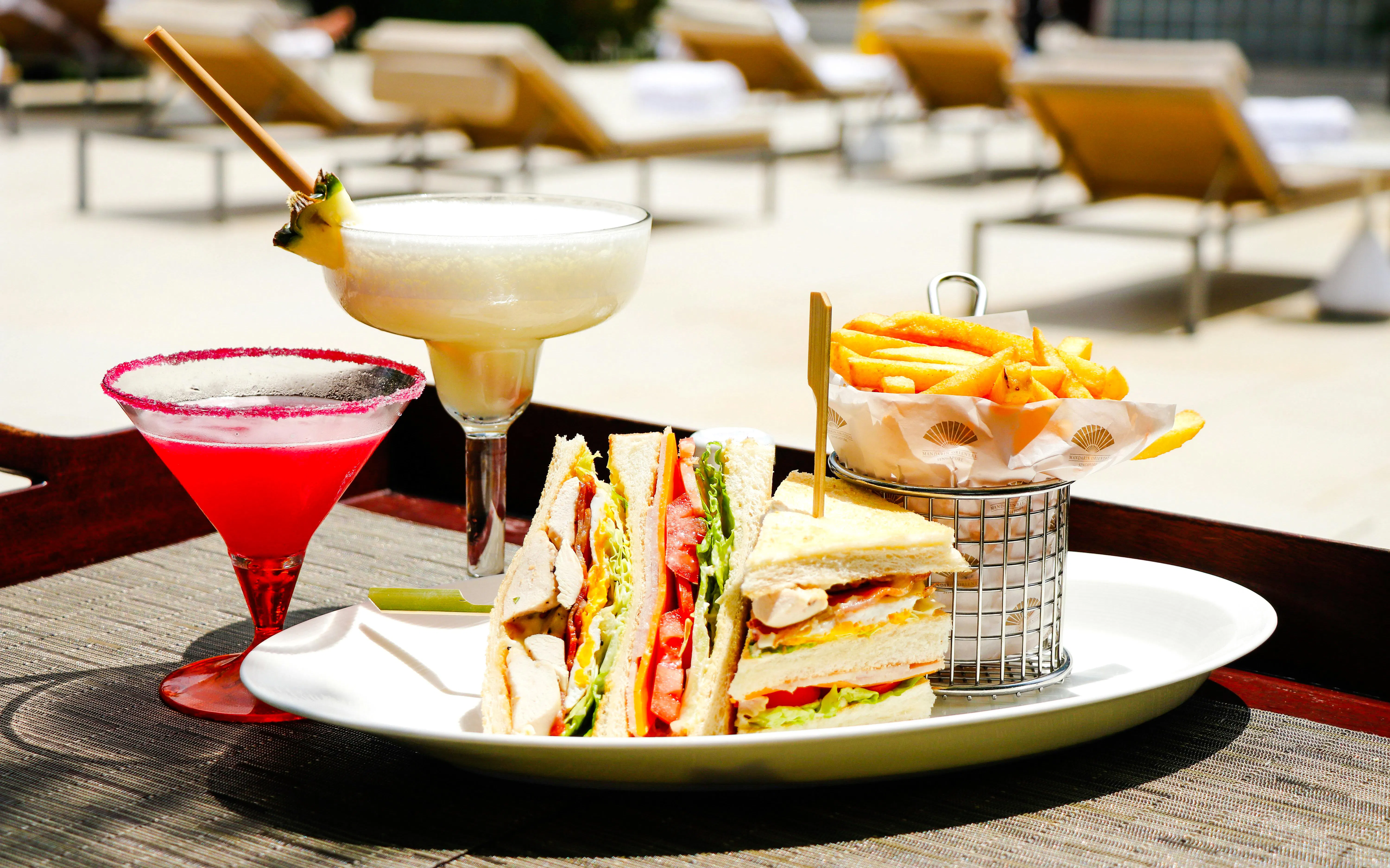 Club sandwich, fries, and cocktails by a poolside lounge.