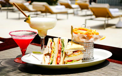 Club sandwich, fries, and cocktails by a poolside lounge.