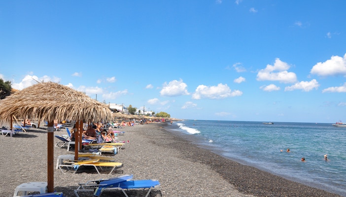Perivolos Beach in Santorini with black sand and umbrella stands along the shoreline.