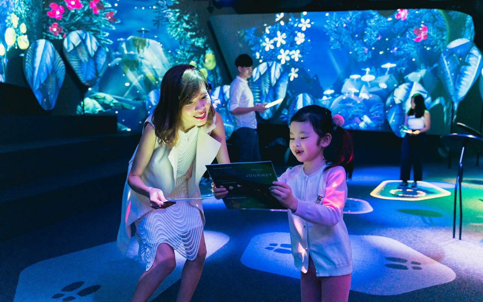 A woman and child with interactive guidebooks, engaged and smiling in the vibrant, digital ambiance of the Changi Experience Studio, Singapore