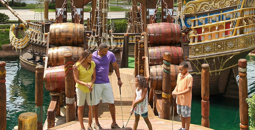 Family playing mini-golf near pirate ship at Pirate's Cove Adventure Golf.