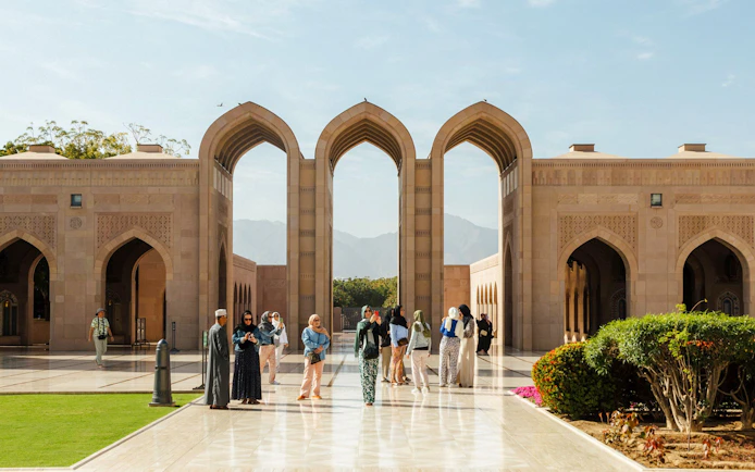 Tourists exploring courtyard of Royal Opera House Muscat with arches and gardens.