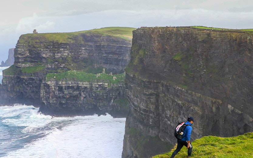 Hiker walking along the Cliffs of Moher in Ireland with ocean waves below.