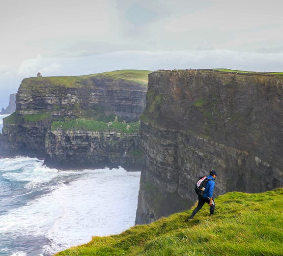 Hiker walking along the Cliffs of Moher in Ireland with ocean waves below.