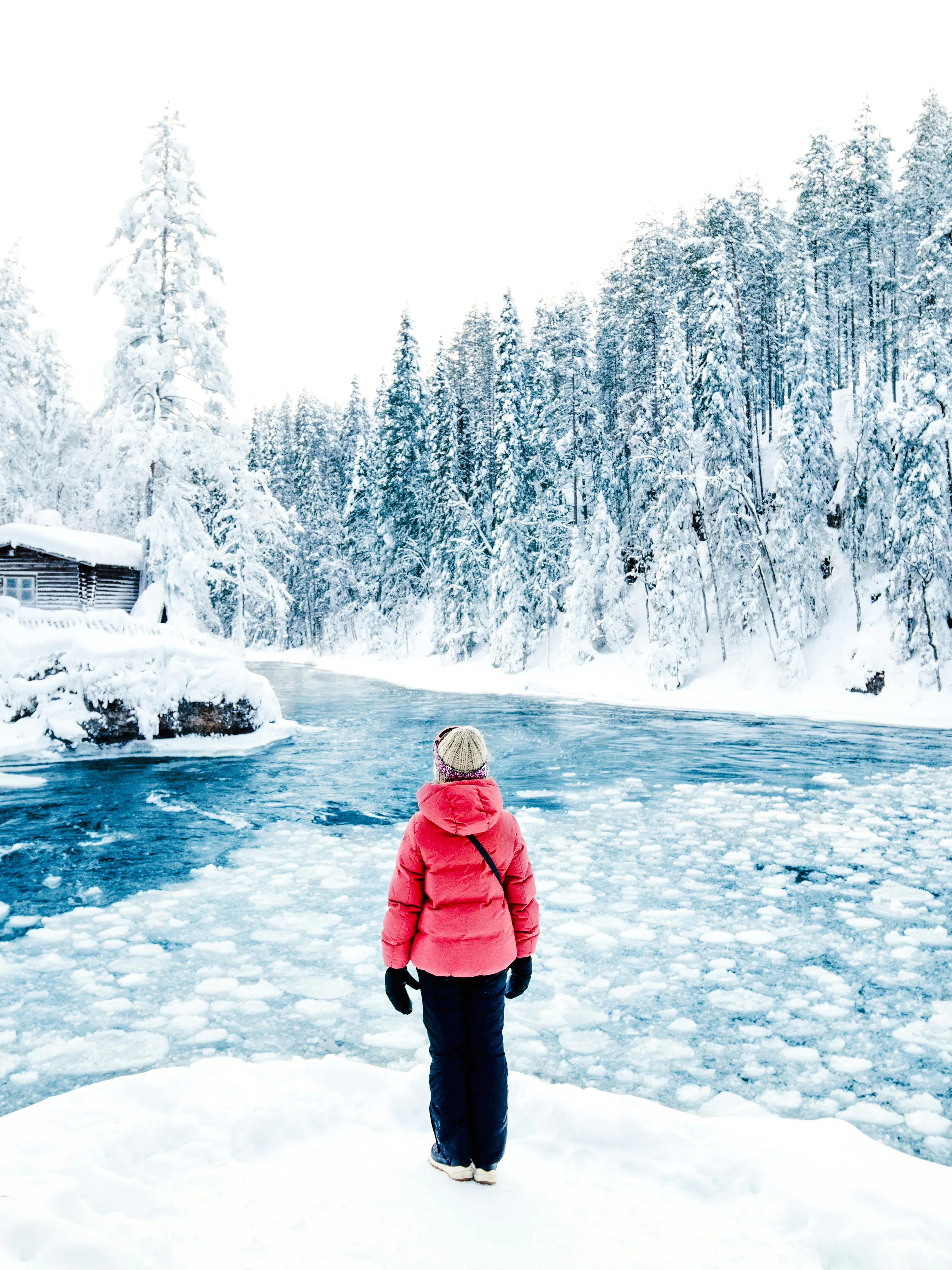 Person standing by a frozen river in Lapland Winter Park, surrounded by snow-covered trees.