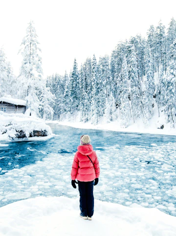 Person standing by a frozen river in Lapland Winter Park, surrounded by snow-covered trees.