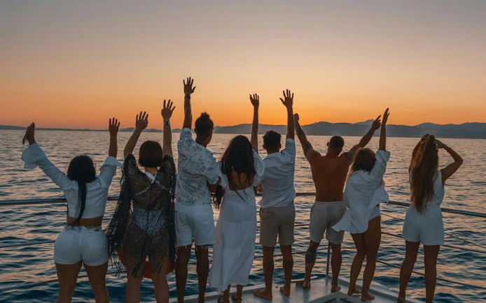 Tourists enjoying a sunset cruise on the ocean, raising hands towards the horizon.