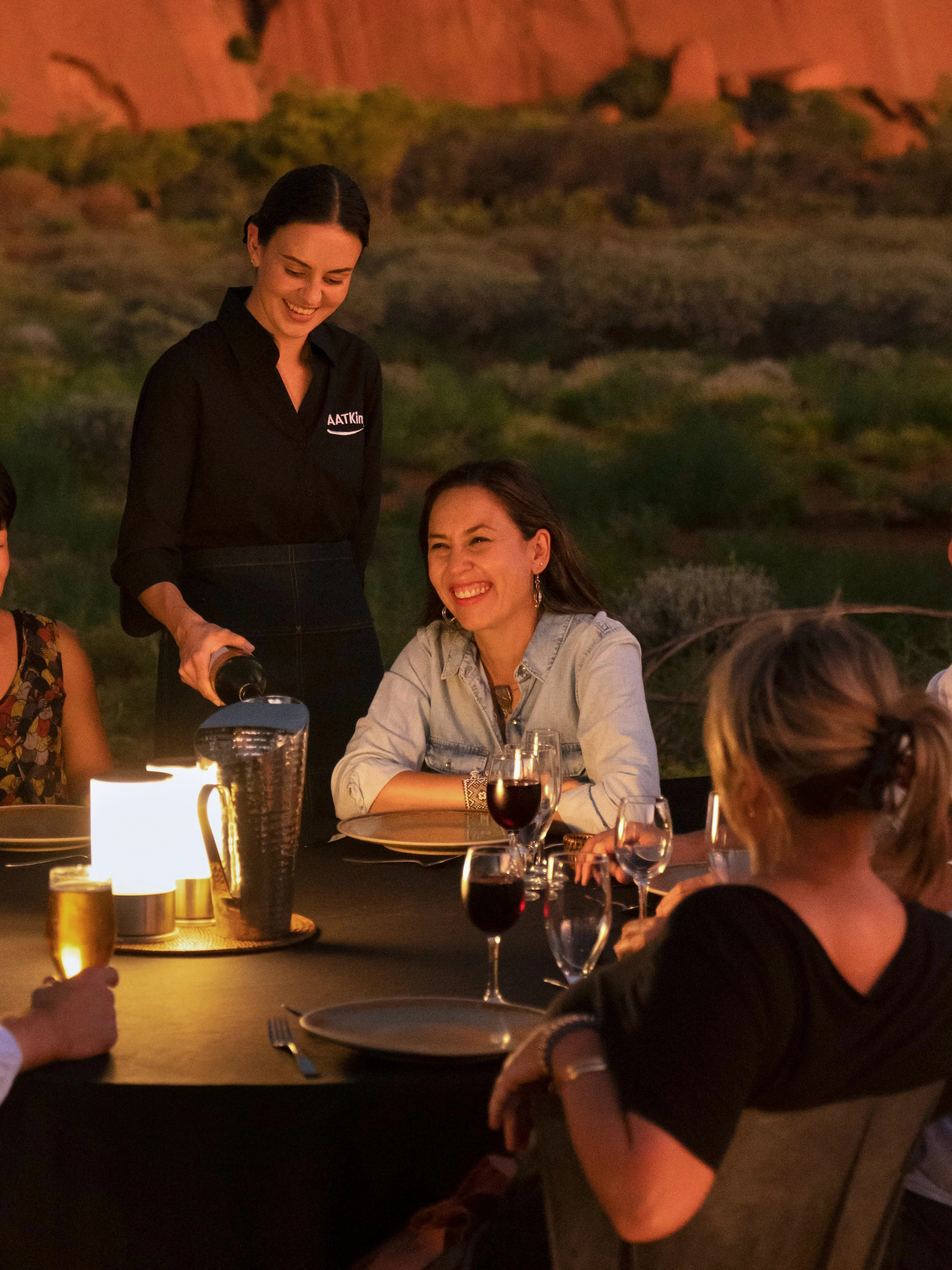 Group dining outdoors at Uluru with a guide serving drinks during sunset.