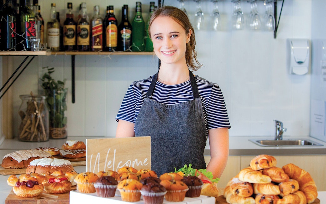 Bakery display with pastries and muffins on a counter, staff member smiling behind.