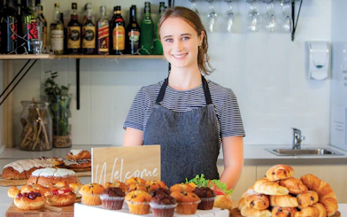 Bakery display with pastries and muffins on a counter, staff member smiling behind.