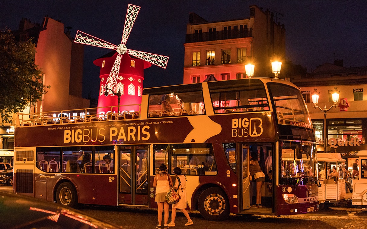 Big Bus Paris at night near illuminated Moulin Rouge.