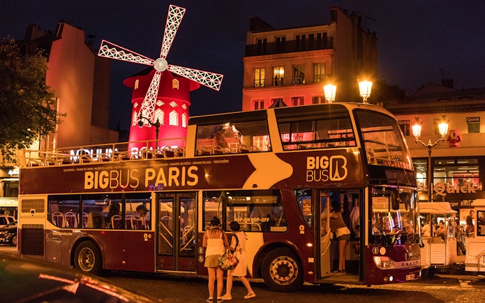 Big Bus Paris at night near illuminated Moulin Rouge.