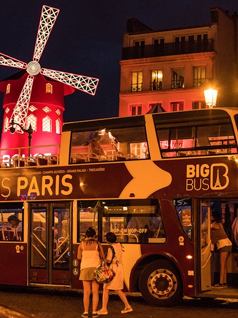 Big Bus Paris at night near illuminated Moulin Rouge.