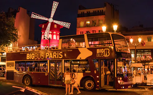 Big Bus Paris at night near illuminated Moulin Rouge.