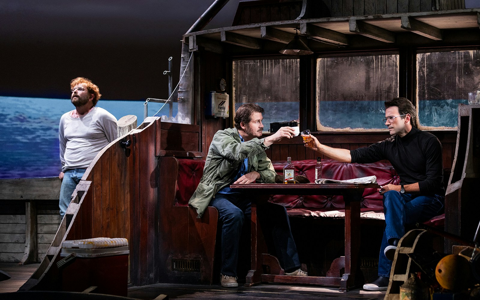 Three men on a boat set, two sharing drinks, in a scene from "The Shark is Broken" play.