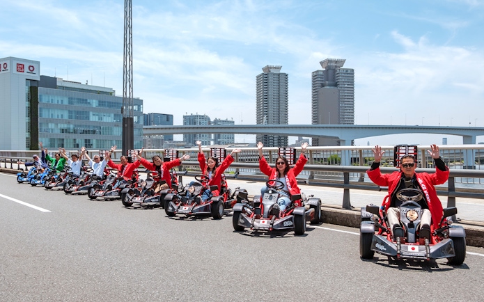 People driving go-karts on Shibuya Shinjuku Tour in Tokyo, with city skyline in background.