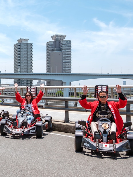 People driving go-karts on Shibuya Shinjuku Tour in Tokyo, with city skyline in background.