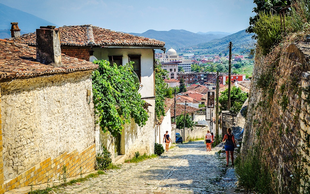 Guests walking along cobblestone street in Berat Old Town, Albania, with historic buildings and distant hills.