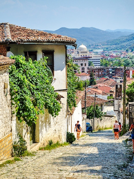 Guests walking along cobblestone street in Berat Old Town, Albania, with historic buildings and distant hills.