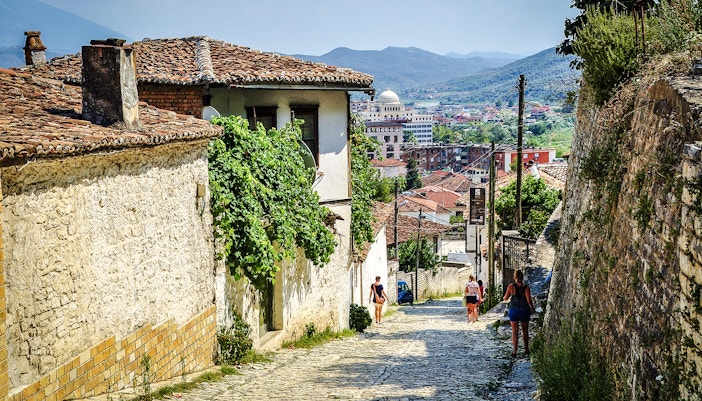 Guests walking along cobblestone street in Berat Old Town, Albania, with historic buildings and distant hills.